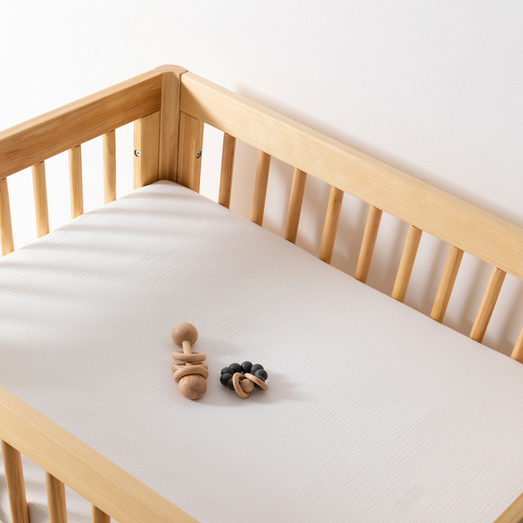 A crib with toy equipped with Babyletto's All-Stages Midi Crib Sheet In GOTS Certified Organic Muslin Cotton in -- Color_White