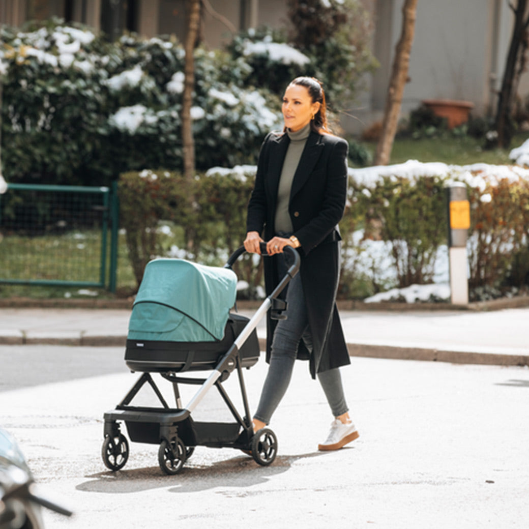 Side view of a young woman taking a walk in the park with her baby in the Thule Shine Stroller in -- Color_Mallard Green