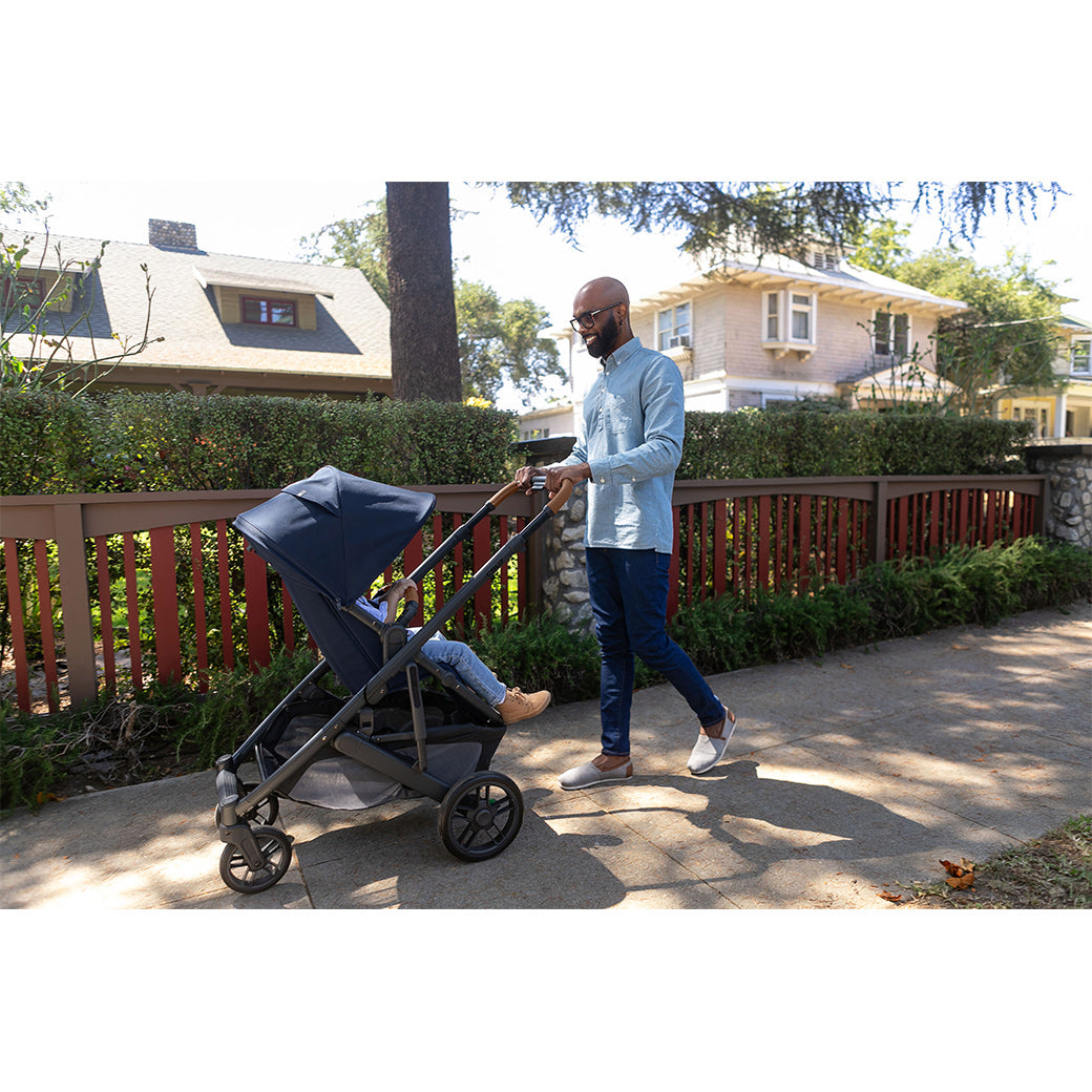 Father with cropped hair, beard and glasses wearing jeans and a long sleeve shirt, smiling at the toddler he is pushing in an uppababy cruz v2 stroller -- Lifestyle
