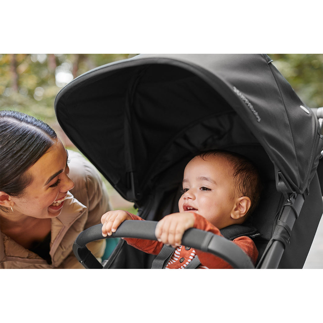Mother with straight black hair wearing a khaki jacket kneeling down smiling at a toddler seated in the uppababy cruz v2 stroller with the sunshade over him -- Lifestyle