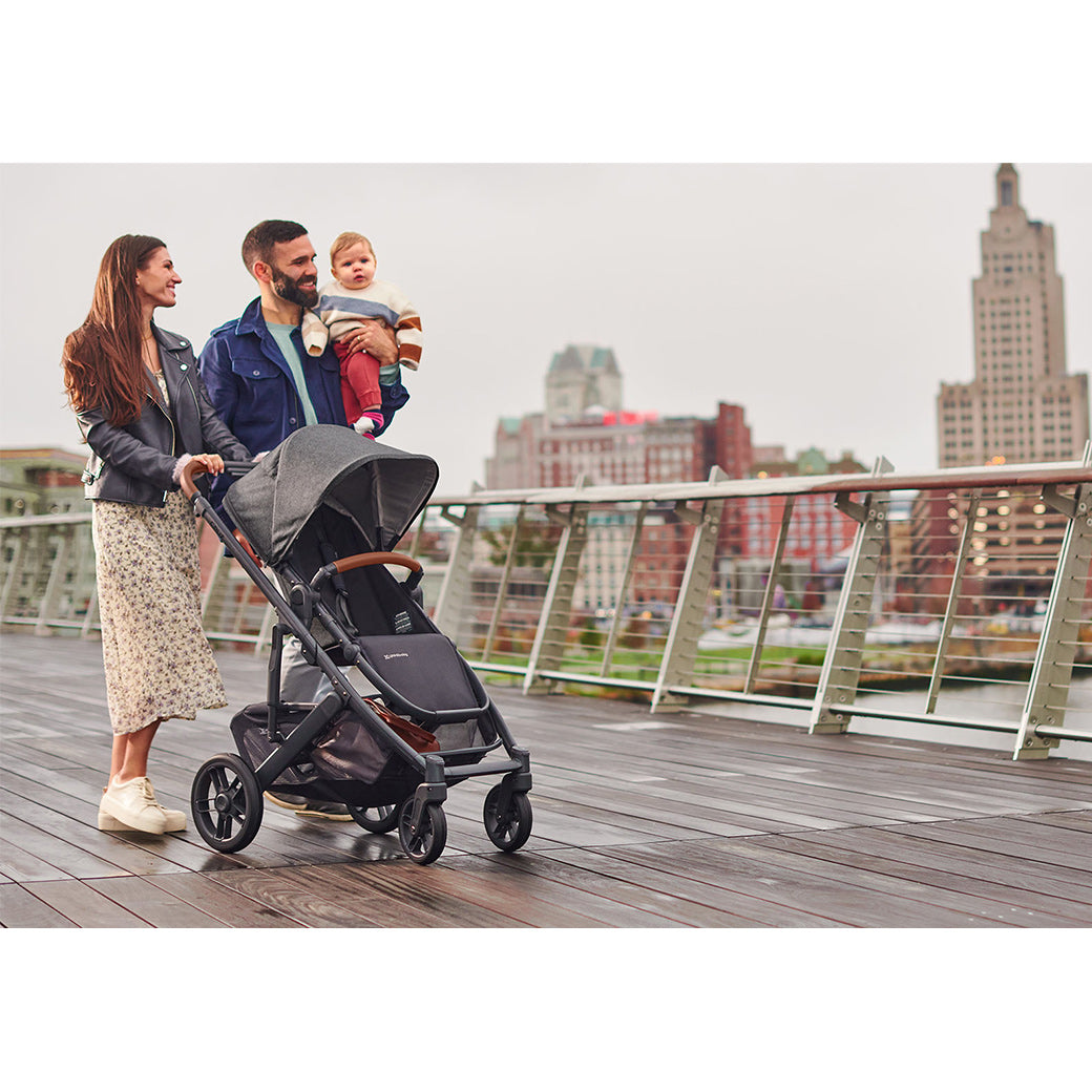 Mother with long brown hair wearing a cream floral skirt, leather jacket and cream sneakers pushing an Uppababy cruz v2 stroller while smiling at father with beard and blue jacket who is walking beside her holding and smiling at baby boy -- Lifestyle