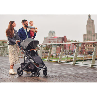 Dad holding the baby and the mom pushing the UPPAbaby CRUZ V2 Stroller in -- Color_Greyson