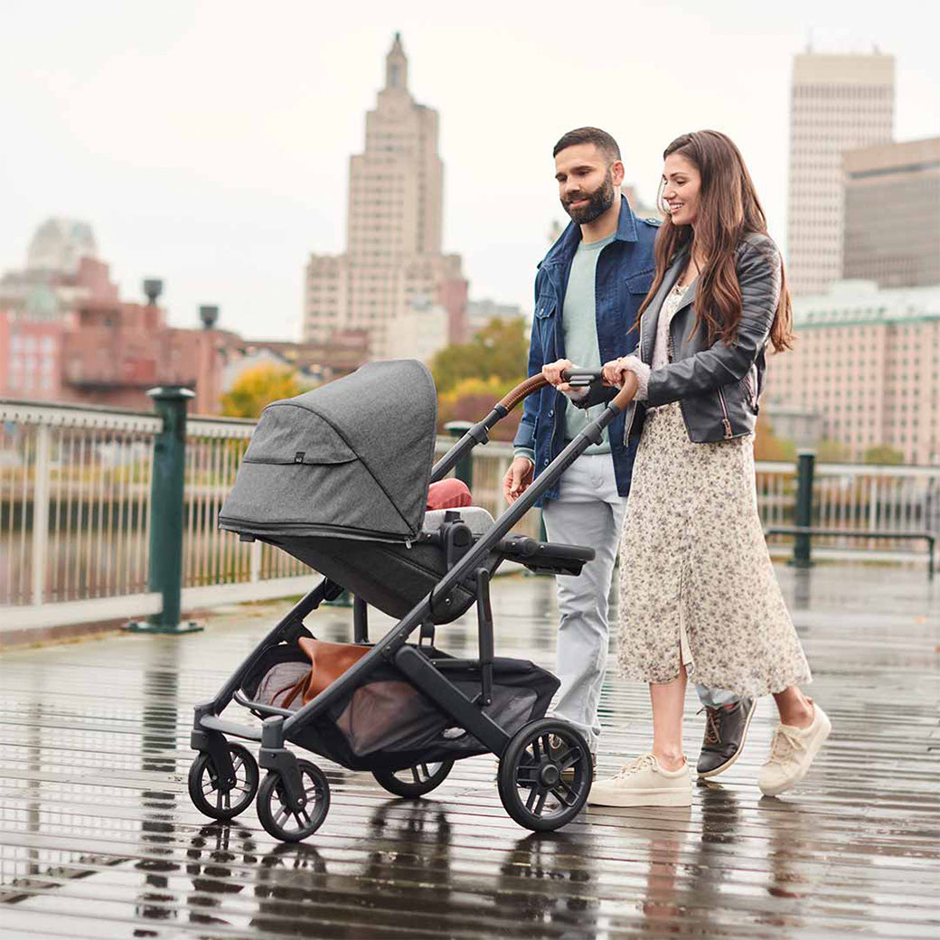 Mother with long brown hair wearing a cream floral skirt, leather jacket and cream sneakers pushing a fully reclined parent-facing Uppababy cruz v2 stroller while she and father smile at reclined baby -- Lifestyle