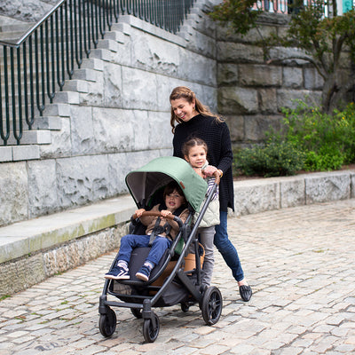 Mom with a toddler pushing a baby down the streen in the UPPAbaby CRUZ V2 Stroller in -- Color_Gwen