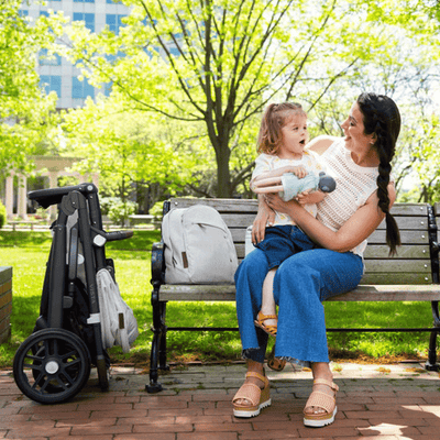 Mommy holding her toddler sitting in a park with the UPPAbaby Changing Backpack on the bench  in - Color_Anthony