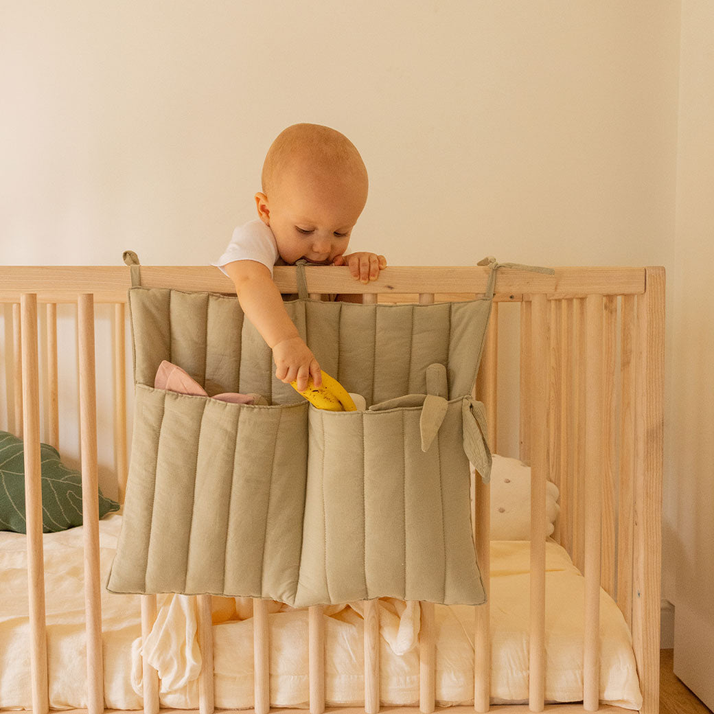 A baby in a crib playing with the Lorena Canals Benjamin Pocket Hanger in -- Color_Olive