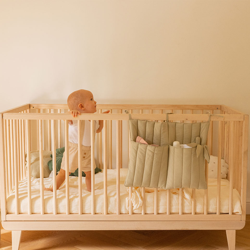 Baby in a crib with Lorena Canals Benjamin Pocket Hanger hanging from the rail  in -- Color_Olive
