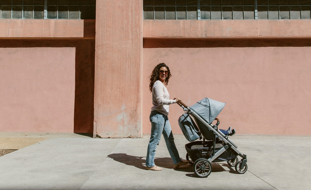 Mother with curly hair wearing jeans and a loose fitting sweater, smiling at the camera while pushing a toddler in the uppababy cruz v2 stroller -- Lifestyle