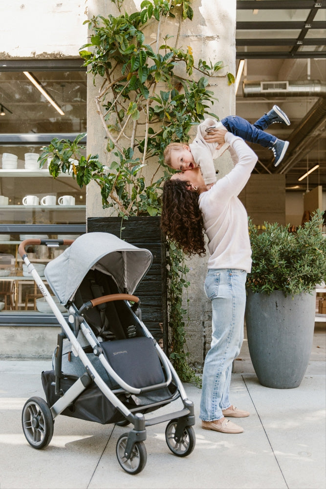 Mother with curly hair wearing jeans and a loose fitting sweater holding toddler child up and kissing his face while he looks at the camera happily, while standing next to the uppababy cruz v2 stroller angling towards the camera -- Lifestyle